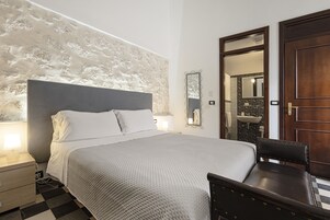 Un dormitorio ordenado con cama, mesita de noche, silla y un baño visible a través de una puerta abierta.