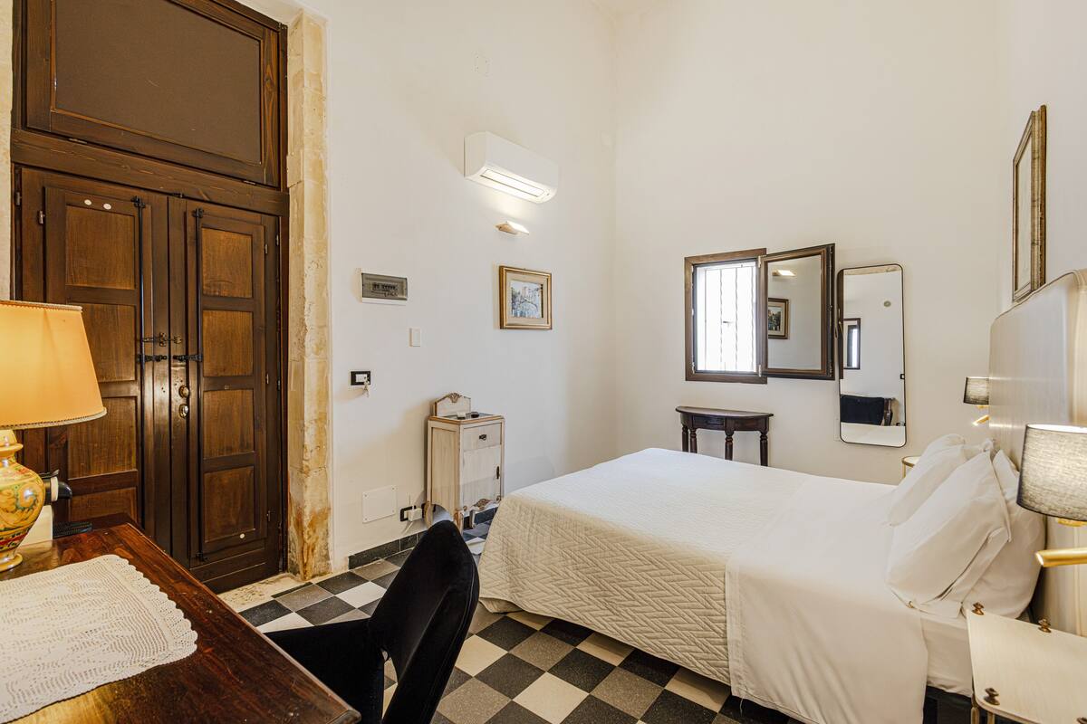 A hotel room with a bed, a desk, a chair, a lamp, a mirror, and a wardrobe.