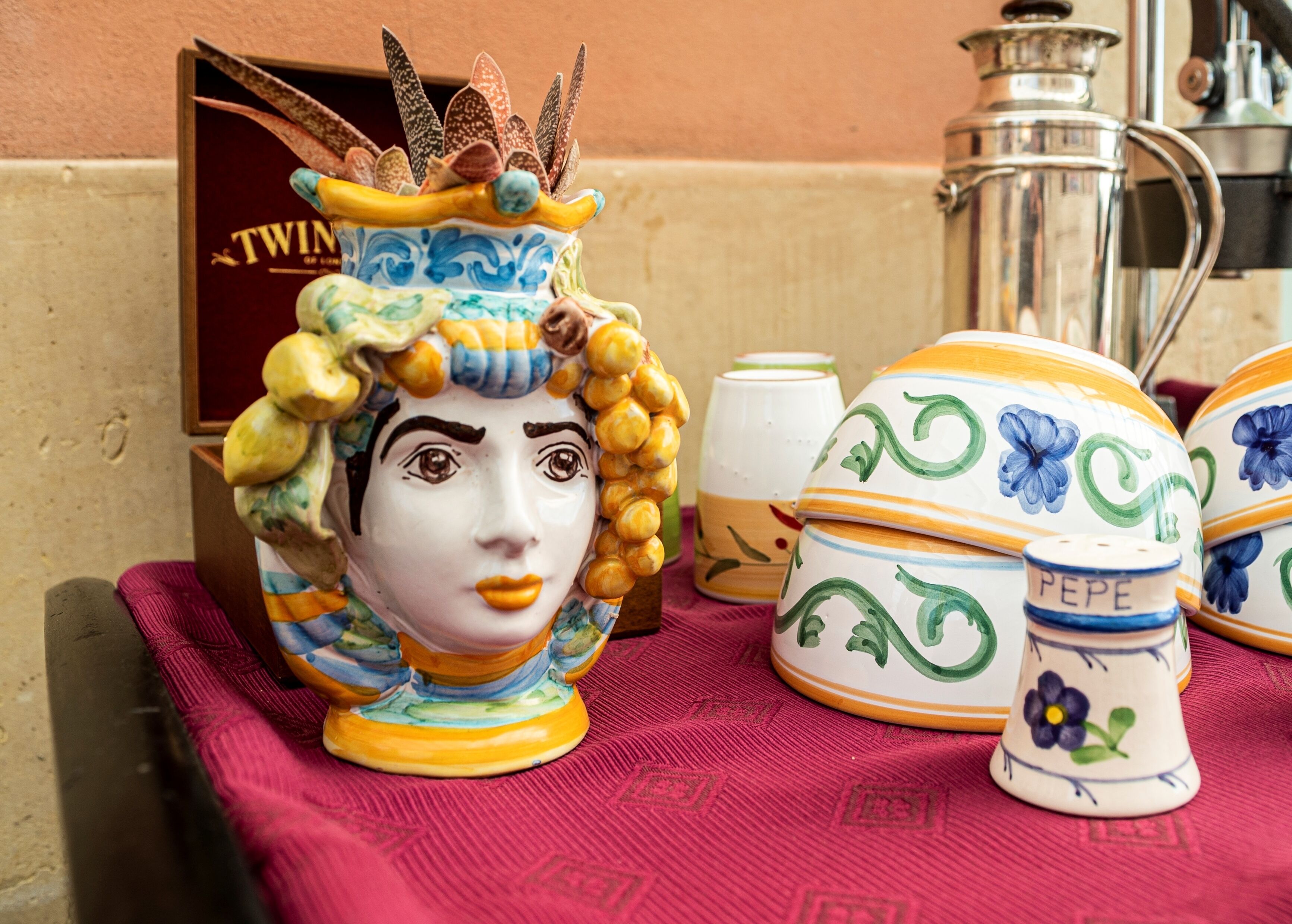 Des objets en céramique sont posés sur une table, notamment un vase orné de motifs floraux et un petit récipient portant l’inscription « PEPE ».