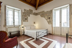 Un dormitorio con pared de piedra, una cama con estructura metálica, una silla roja con estampado y una alfombra blanca.