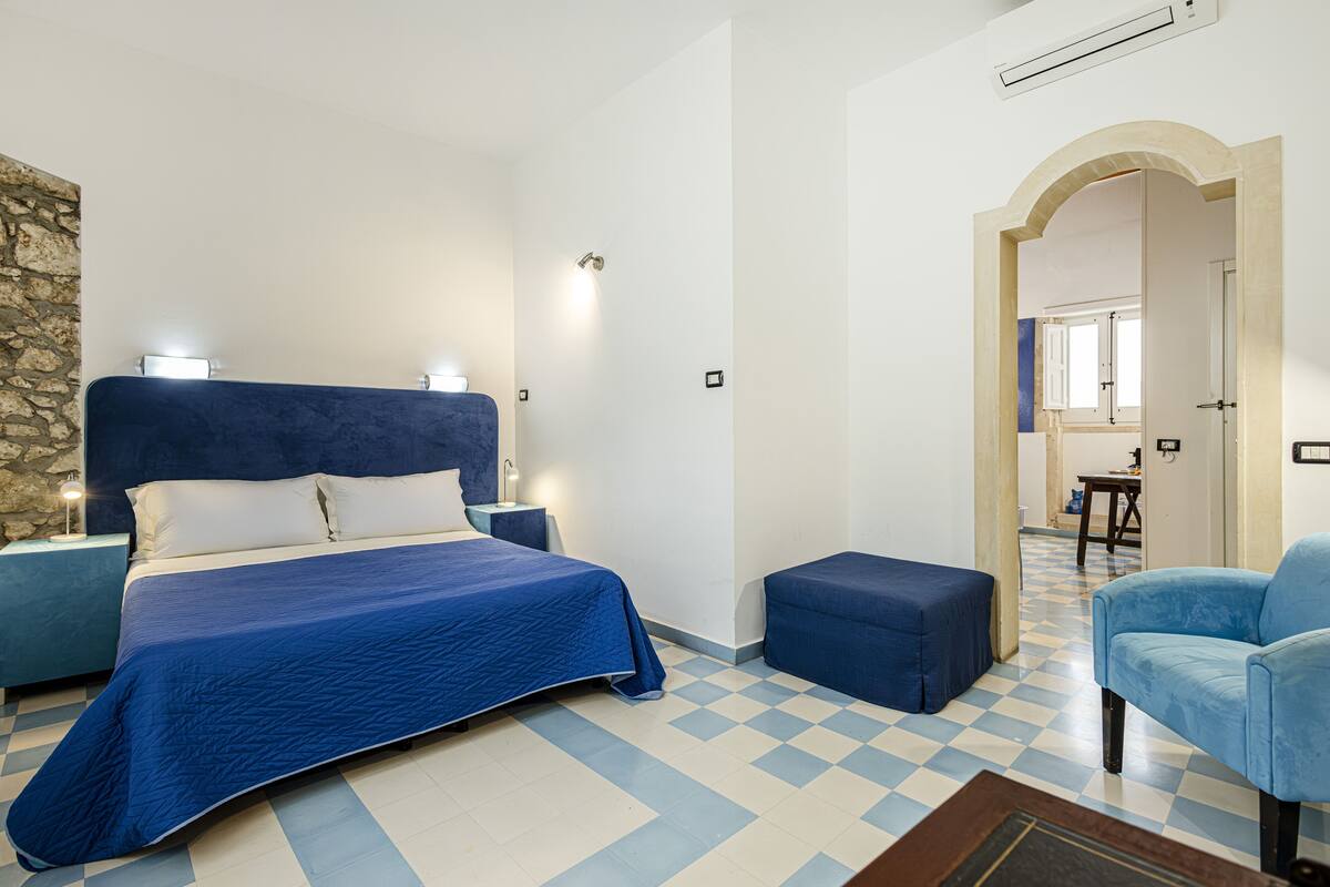 A bedroom with a large bed, a blue chair, a small table, and a checkered floor.