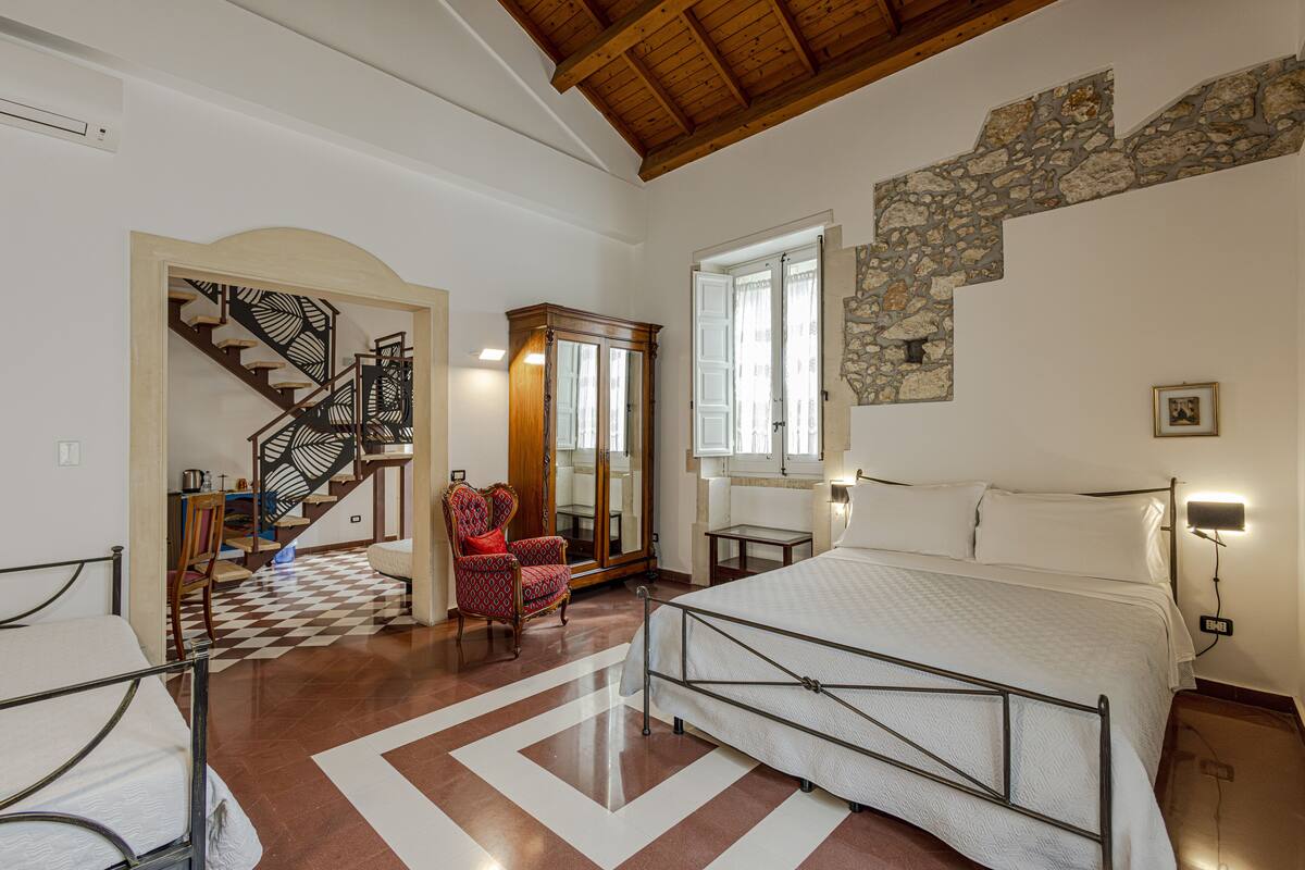 A bedroom with a large bed, a chair, a desk, a mirror, and a staircase leading to another room.