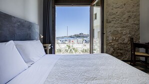 Ein Hotelzimmer mit einem großen Bett, Blick auf die Stadt durch eine offene Tür, einer Steinsäule und einem Balkon mit Geländer.