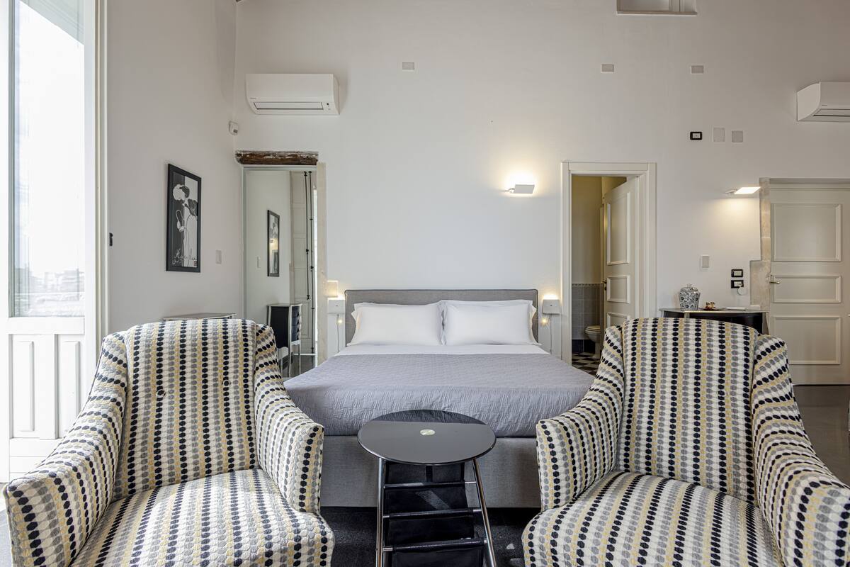 A hotel room with a bed, two patterned armchairs, a small table, and a mirror.