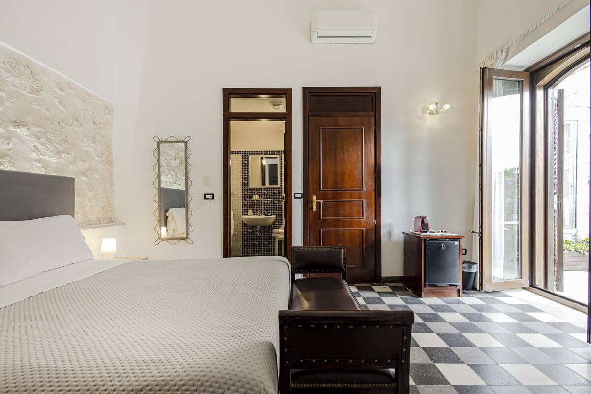 A hotel room with a bed, a desk, a chair, a mirror, a sink, and a view through a glass door.