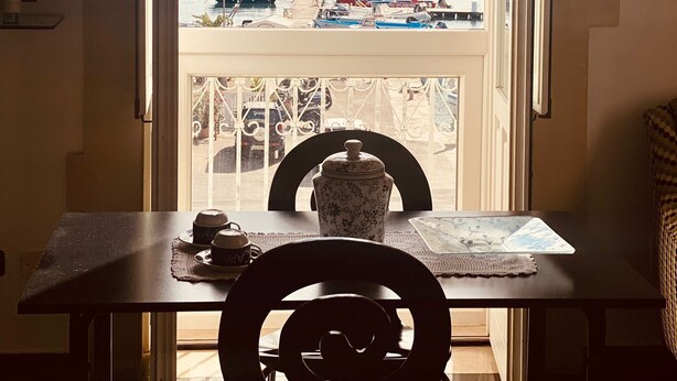 Ein Zimmer mit Tisch und Stühlen, Blick auf einen Hafen mit Booten und Gebäuden, und einem klaren Himmel.