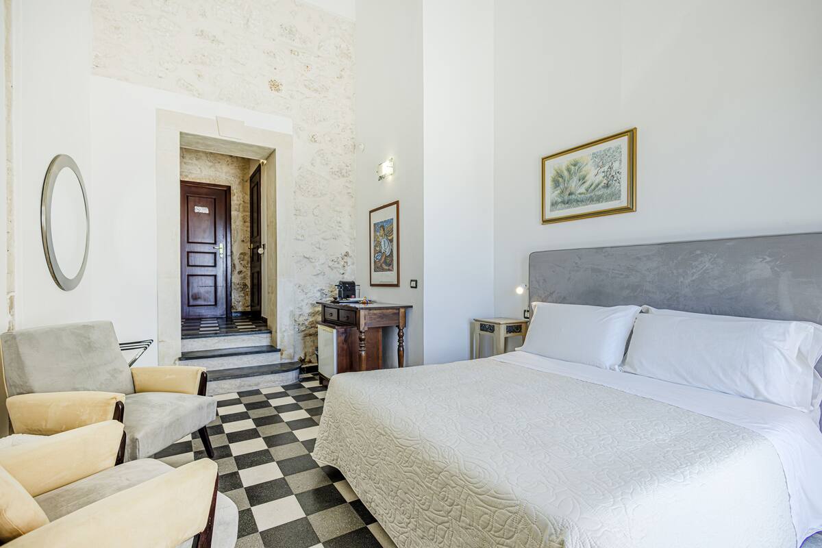 A hotel room with a checkered floor, a bed with white linens, a desk with a chair, a mirror, and a framed picture on the wall.