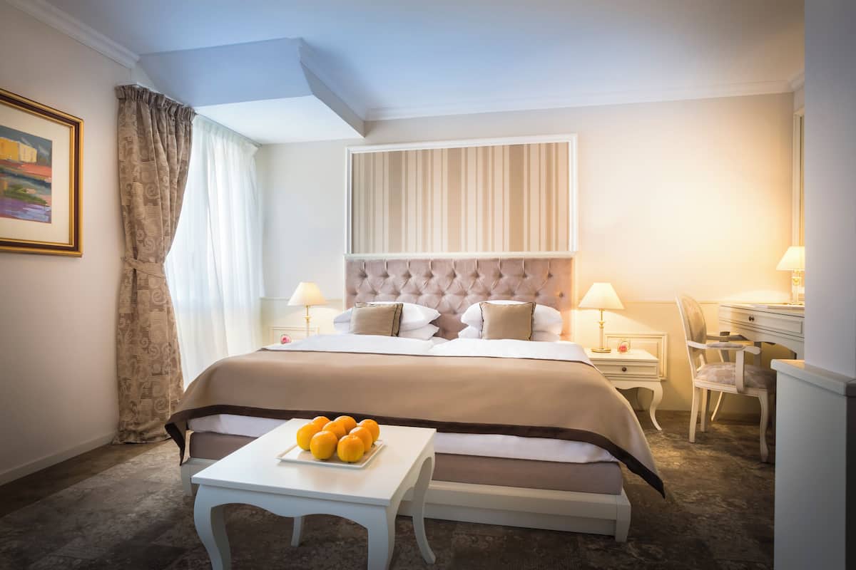 A hotel room with a large bed, a bedside table with a lamp, a chair, and a small table with oranges.