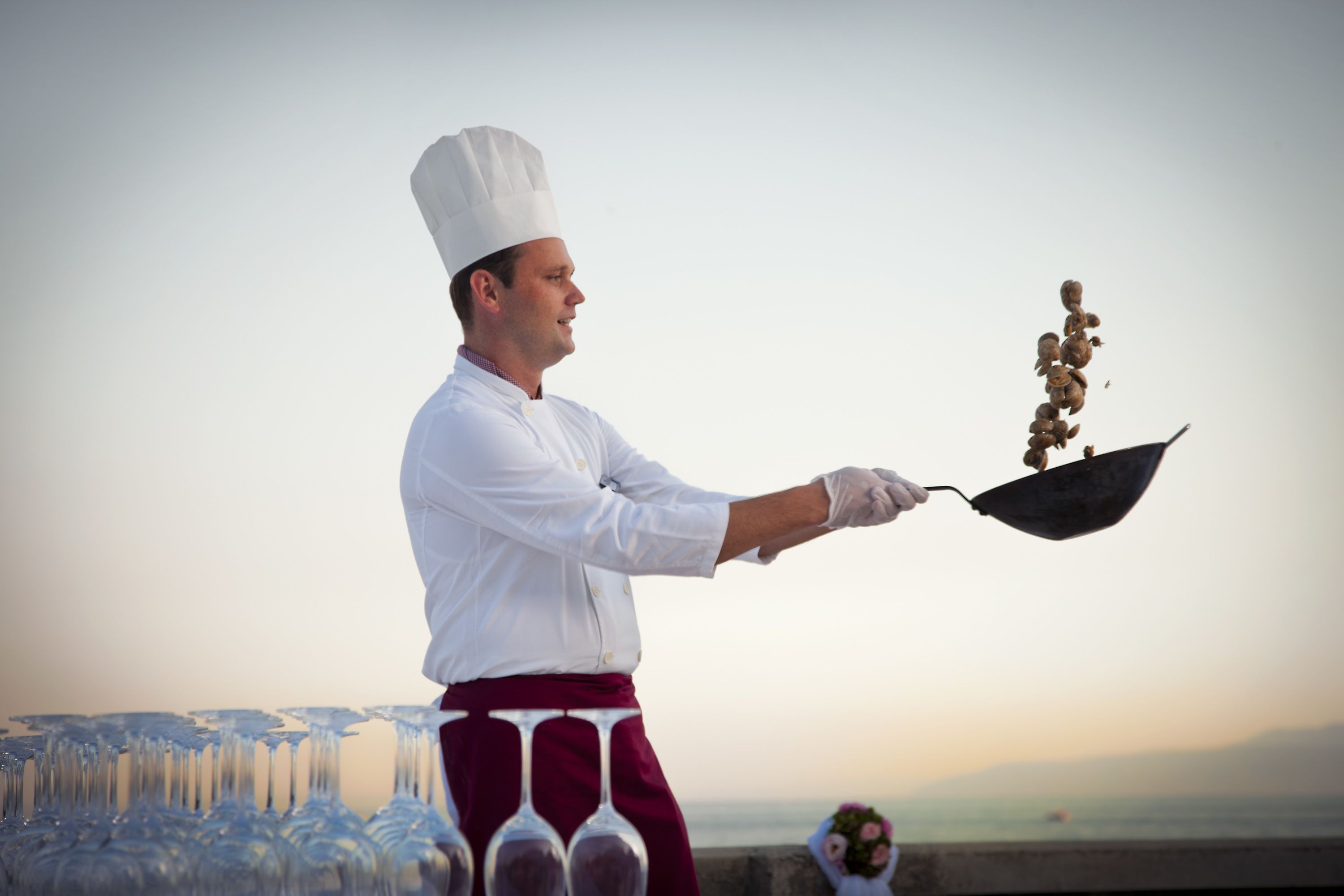 A chef holding a wok with food, standing in front of a row of wine glasses and a bouquet of flowers.