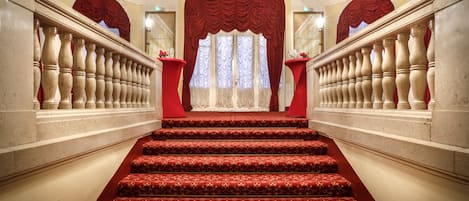 Una imponente escalera con peldaños alfombrados en rojo y barandillas ornamentales, que conduce a un gran arco de entrada con cortinas rojas.