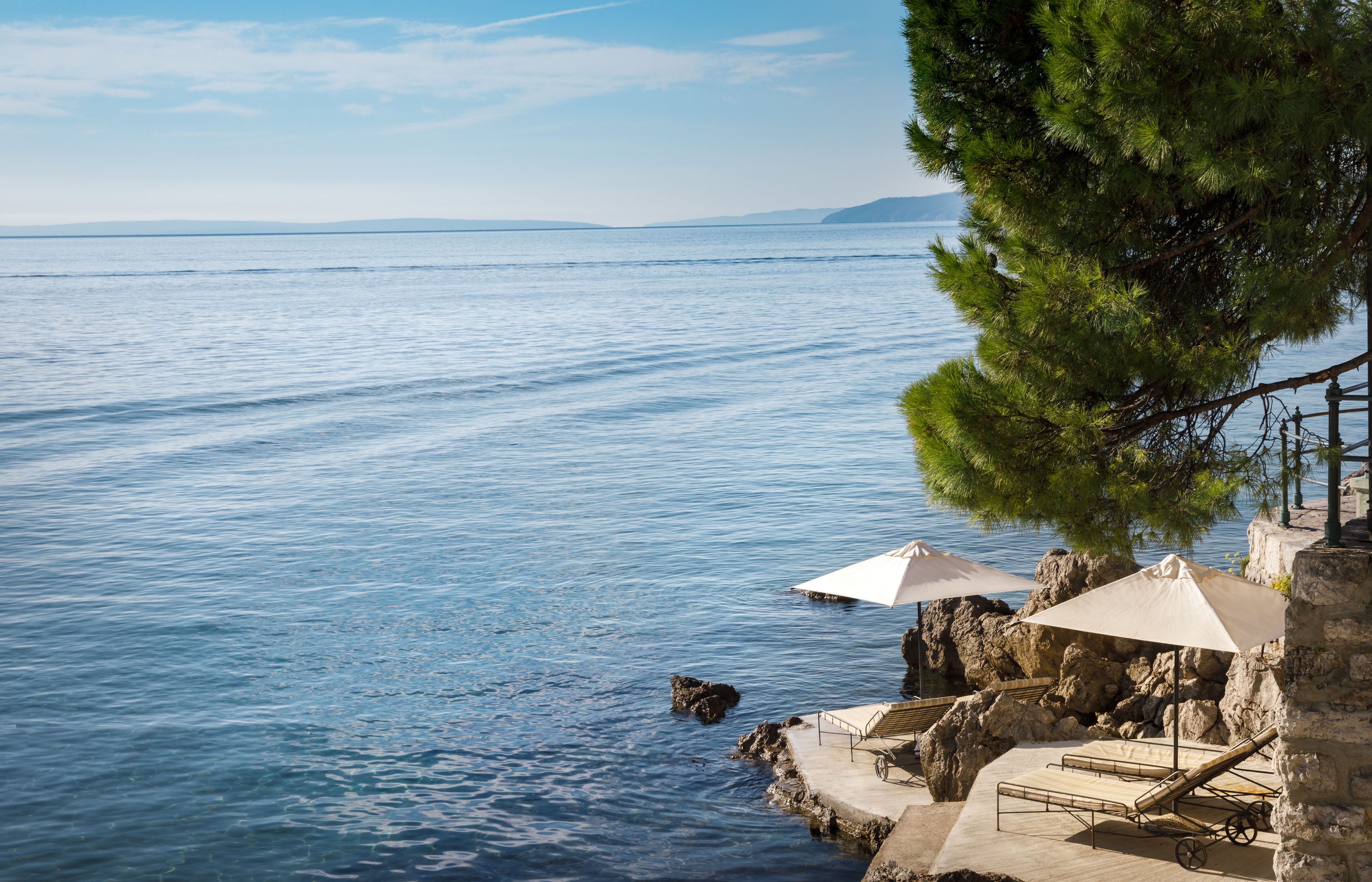 A coastal view with a clear sky, calm sea, and a stone structure with outdoor seating and umbrellas.