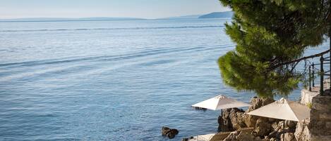 A coastal view with a clear sky, calm sea, and a stone structure with outdoor seating and umbrellas.