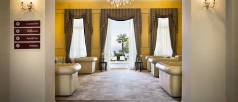 A luxurious hotel lobby with a grand chandelier, two plush sofas, a carpeted floor, and a view of a pool through large windows.