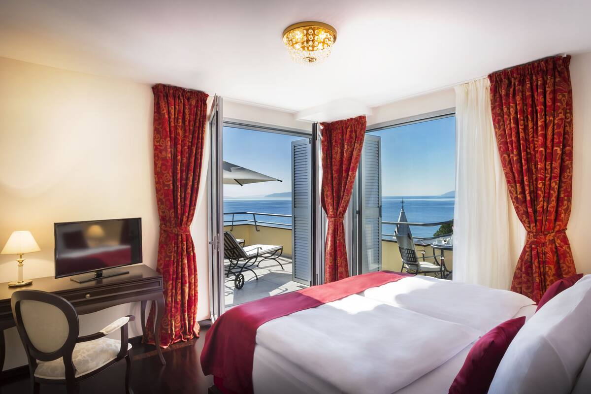 A hotel room with a bed, a desk with a TV, a chair, and a balcony with a table and chairs.