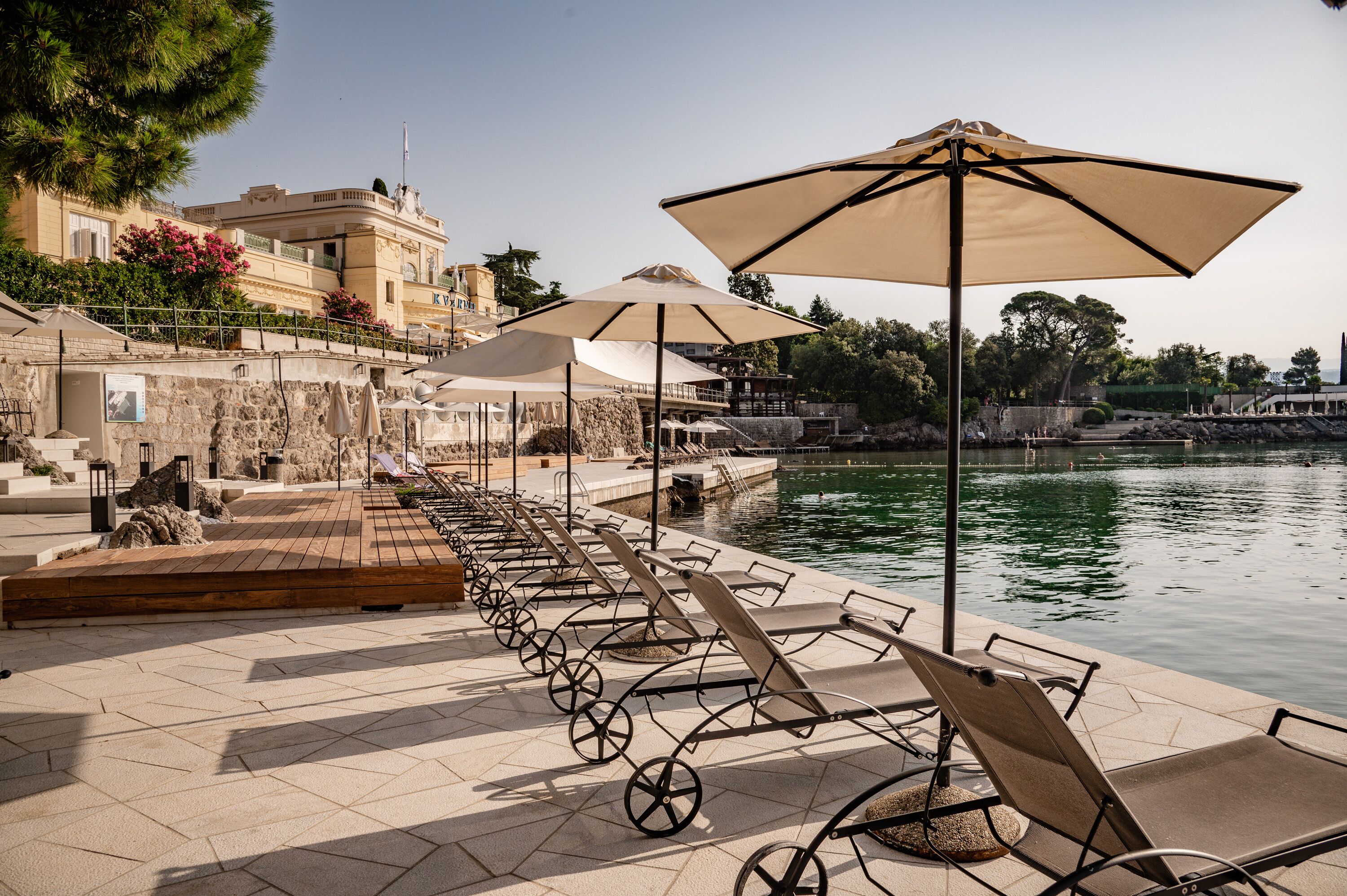 A waterfront terrace with sun loungers and umbrellas, a stone pathway, and a view of the water and buildings in the background.