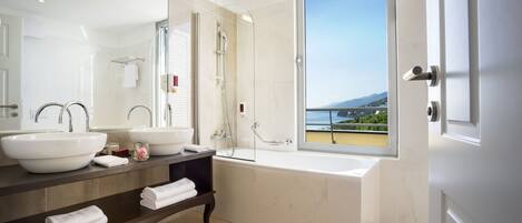 A modern bathroom with two sinks, a large mirror, a bathtub, and a view of the sea through a window.