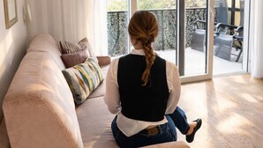 A woman sitting on a beige sofa in a room with a large sliding glass door leading to an outdoor area with chairs. - Gartenresidence Zea Curtis (Merano)