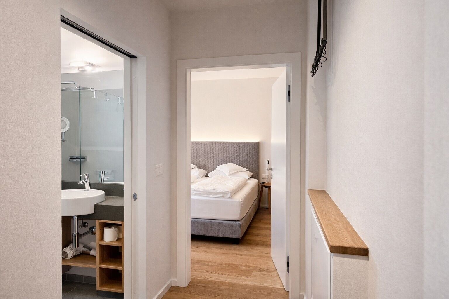Un couloir étroit mène à une chambre qui contient un lit, une table de chevet et une commode. Une salle de bain avec un lavabo et un miroir est visible sur la gauche.