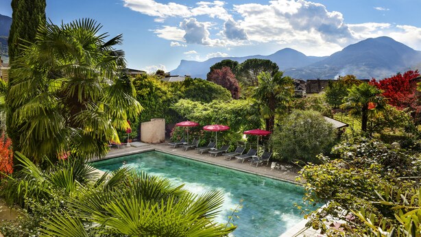 A pool area with lounge chairs and umbrellas, surrounded by lush greenery and mountains in the background. - Gartenresidence Zea Curtis (Merano)