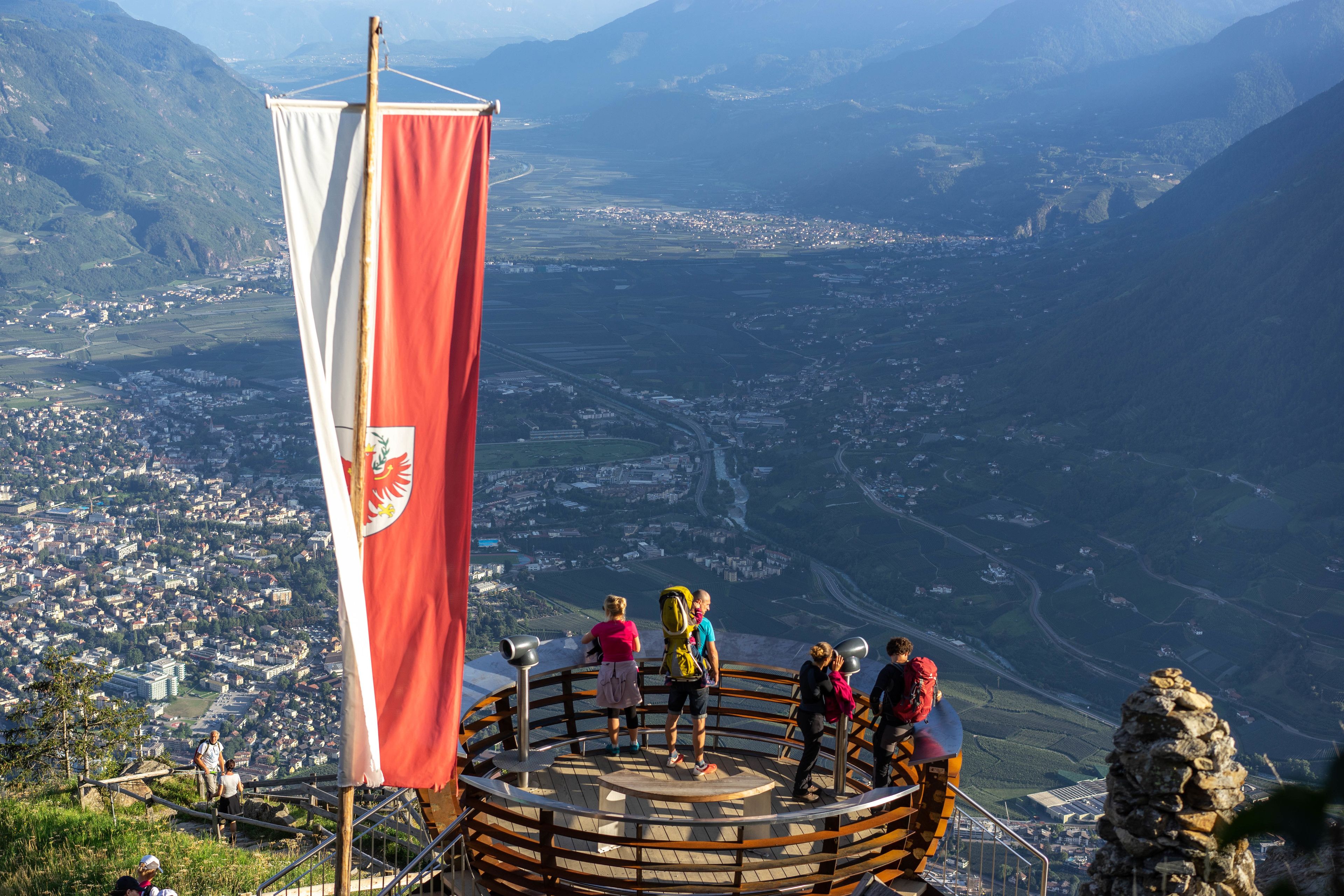 Eine runde Aussichtsplattform mit einer Flagge, die über ein Tal mit einer Stadt und Bergen blickt.