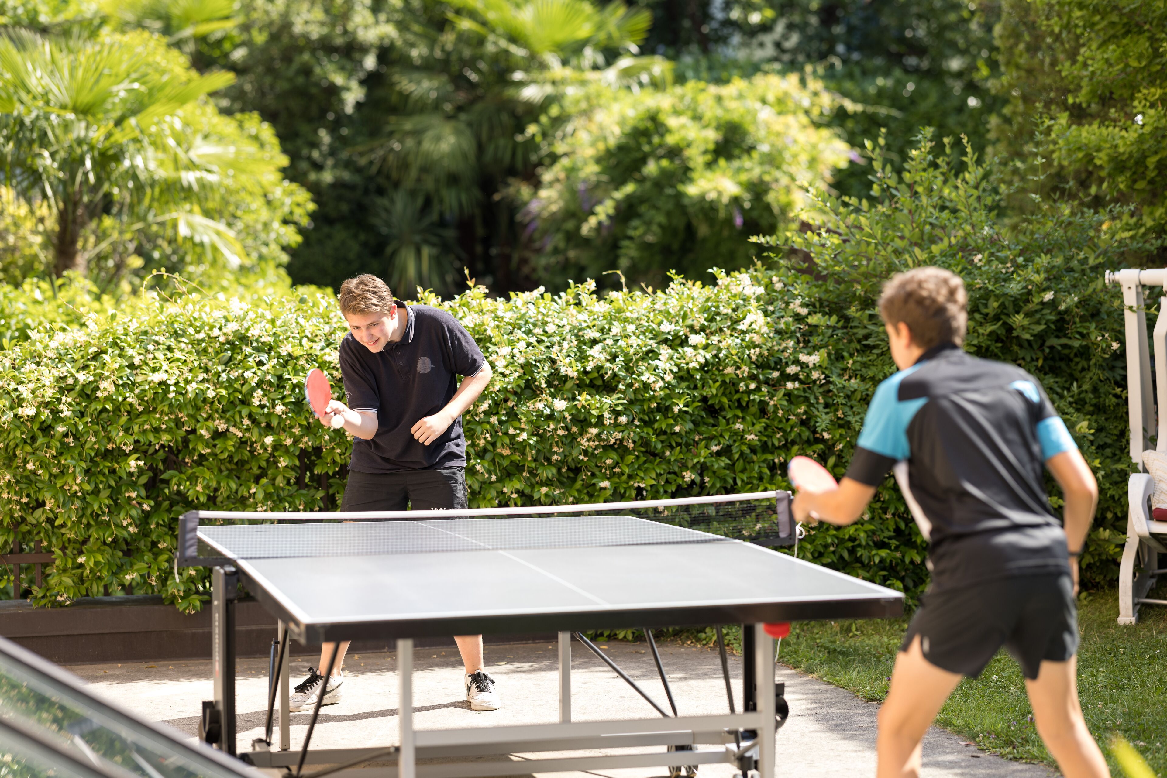 Two people playing table tennis outdoors with a green hedge in the background.