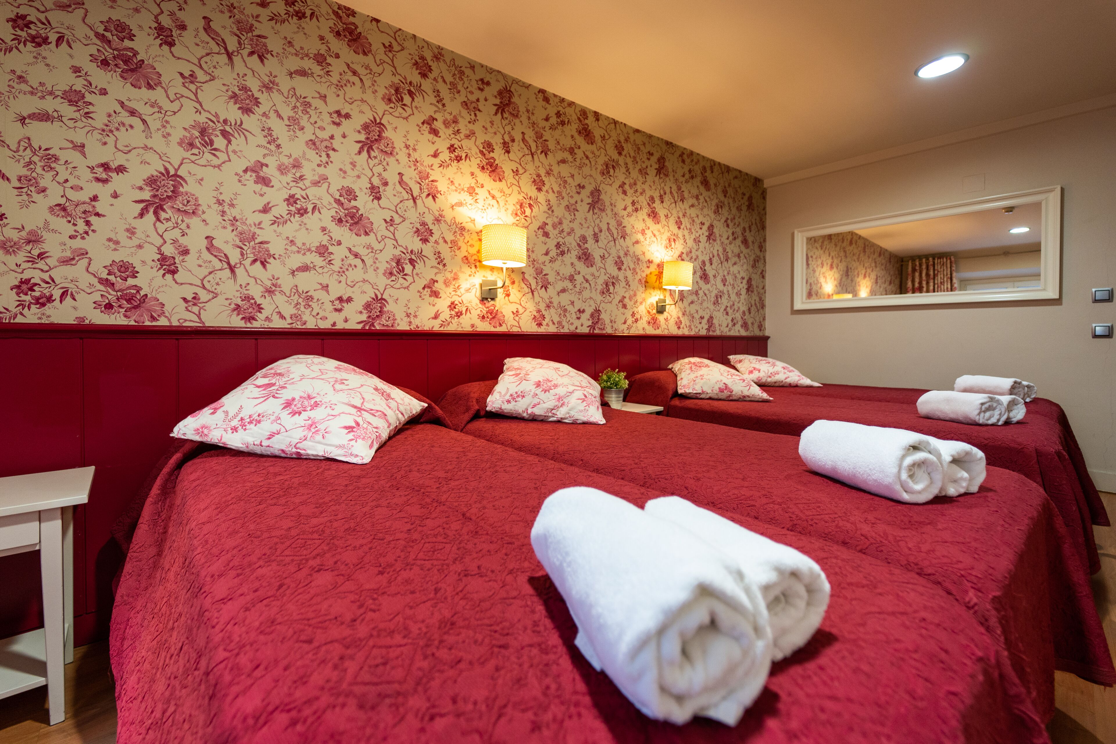 A hotel room with two beds, red headboards, floral wallpaper, and two wall-mounted lamps.