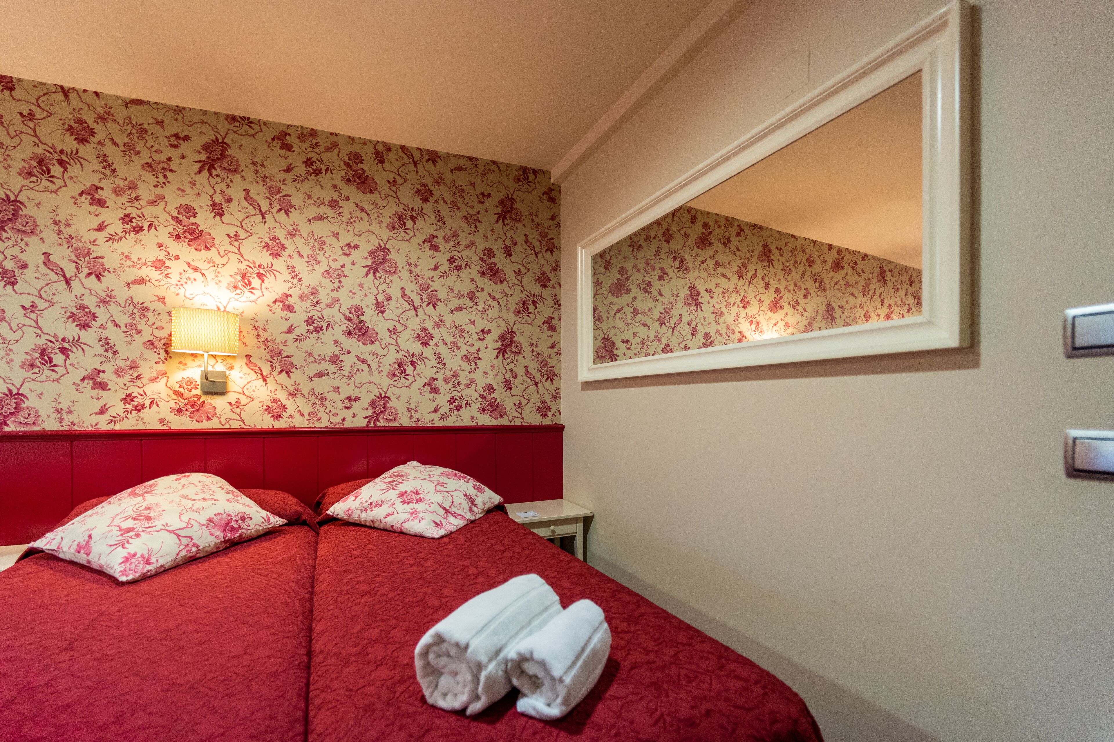 A hotel room with floral wallpaper, a red headboard, a bedside lamp, a mirror, and a neatly made bed with towels.