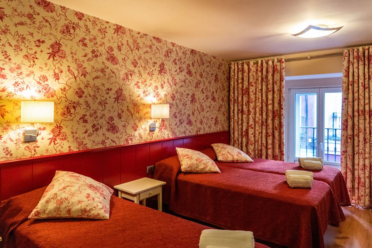 Hotellihuone, jonka seinillä on kukkatapetti, kaksi samanlaisia tyynyjä sisältävää sänkyä, pieni pöytä ja ikkuna, jossa on verhot.