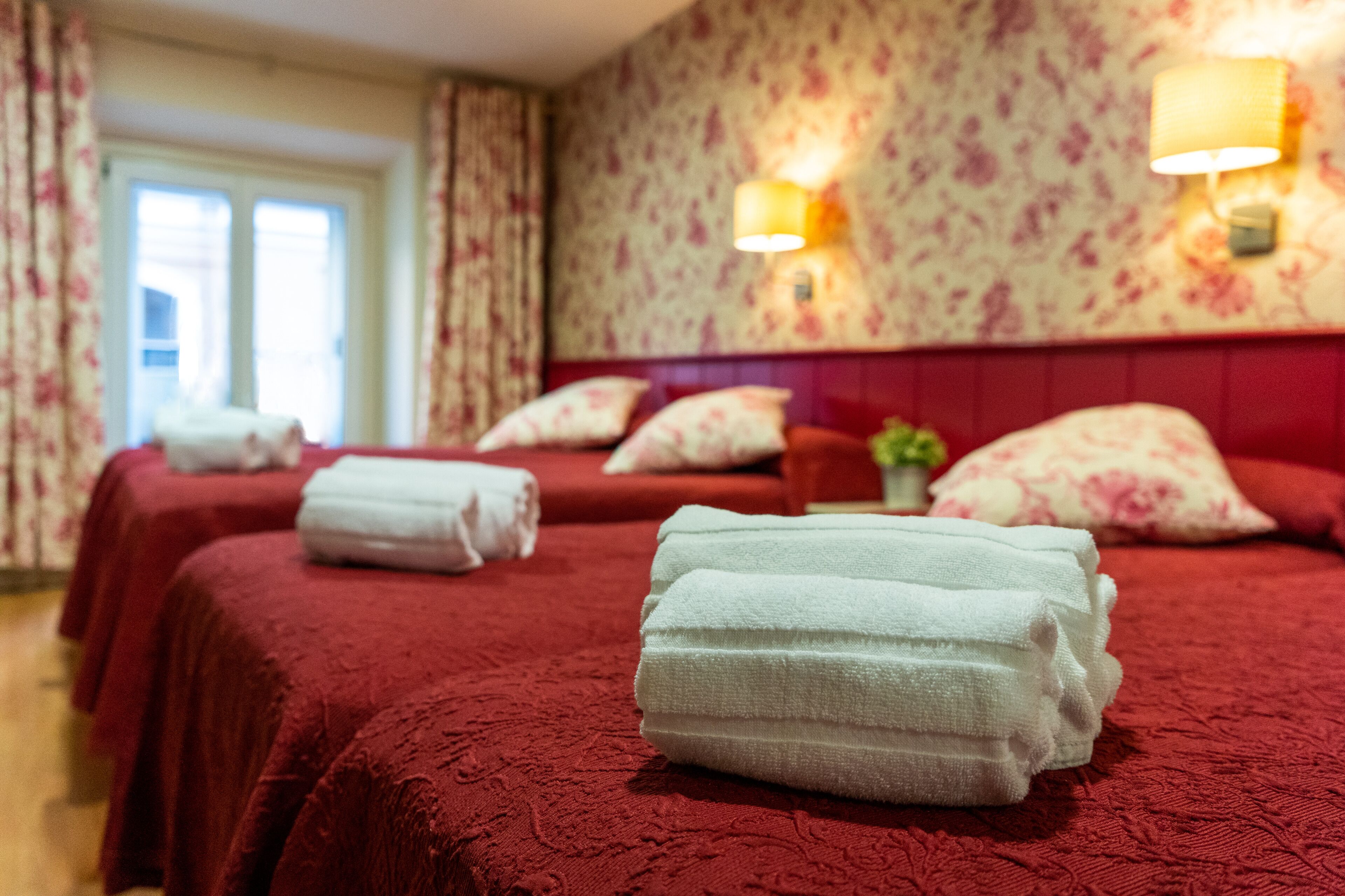 A hotel room with two beds, each with a red bedspread and white towels. There are floral-patterned curtains and wall sconces with lamps.