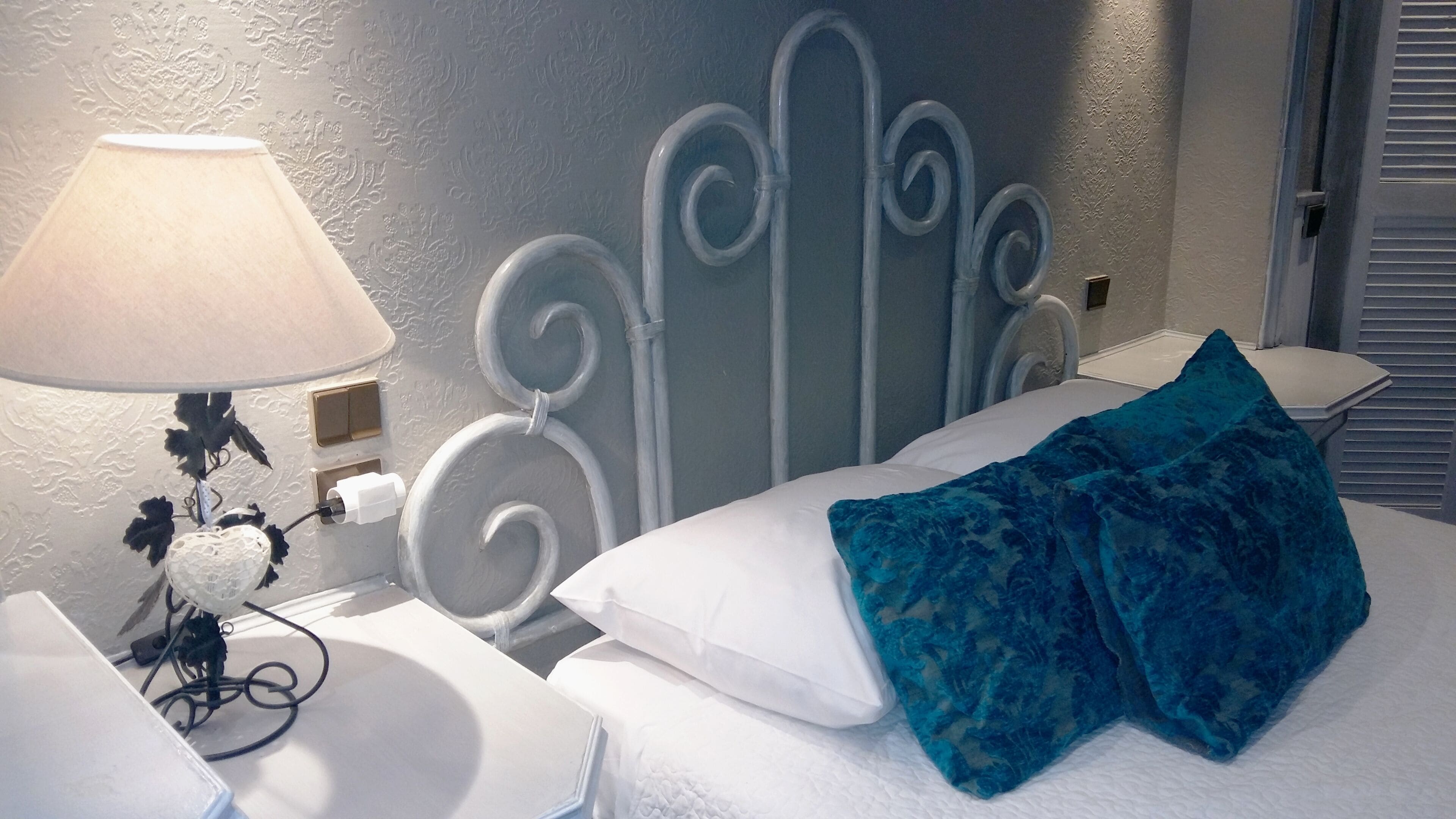 A neatly made bed with white linens and blue pillows, a bedside table with a lamp, and a wall with a decorative metal headboard.