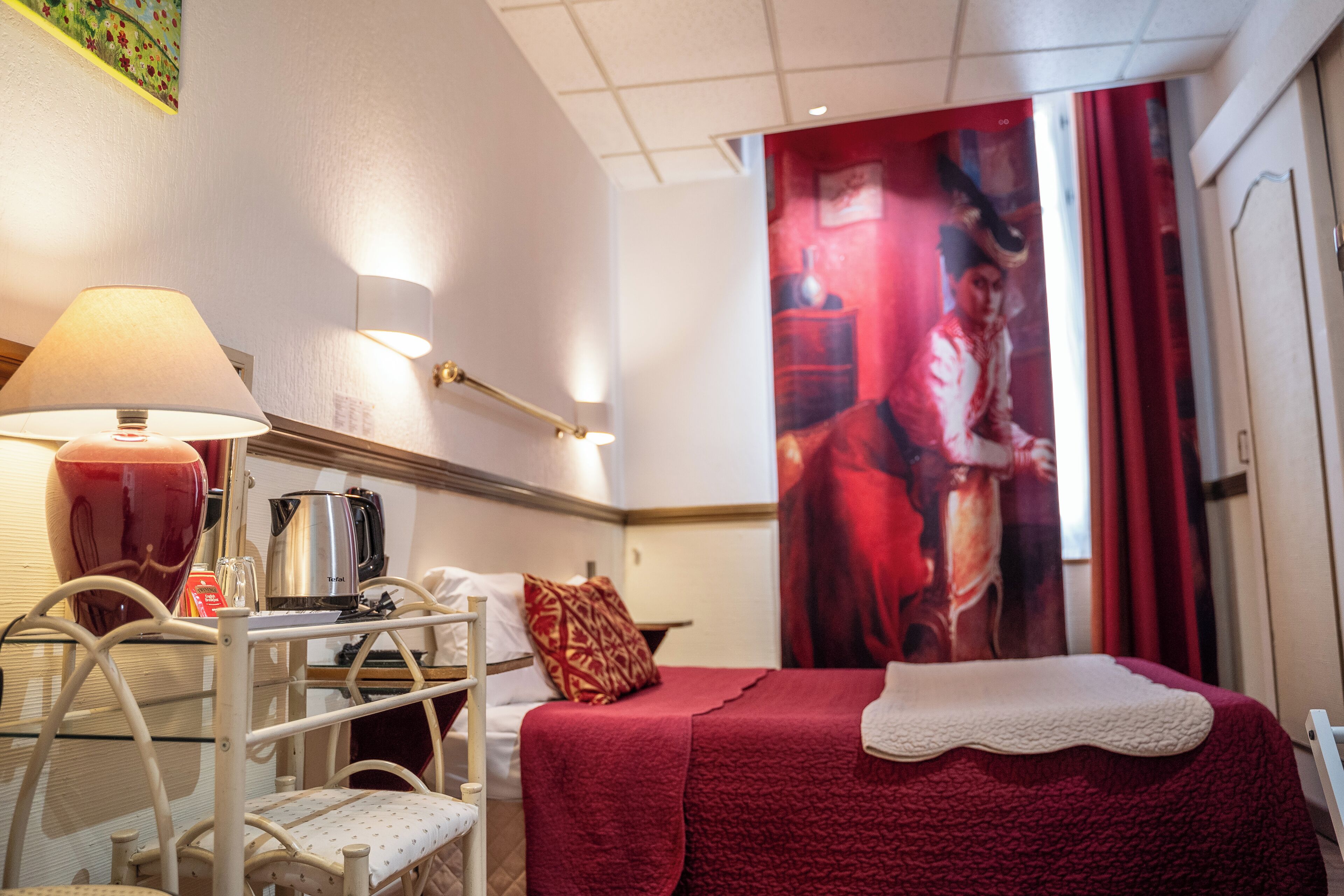 Una camera d'albergo con un letto, una scrivania con una lampada, una sedia e un grande quadro appeso al muro.