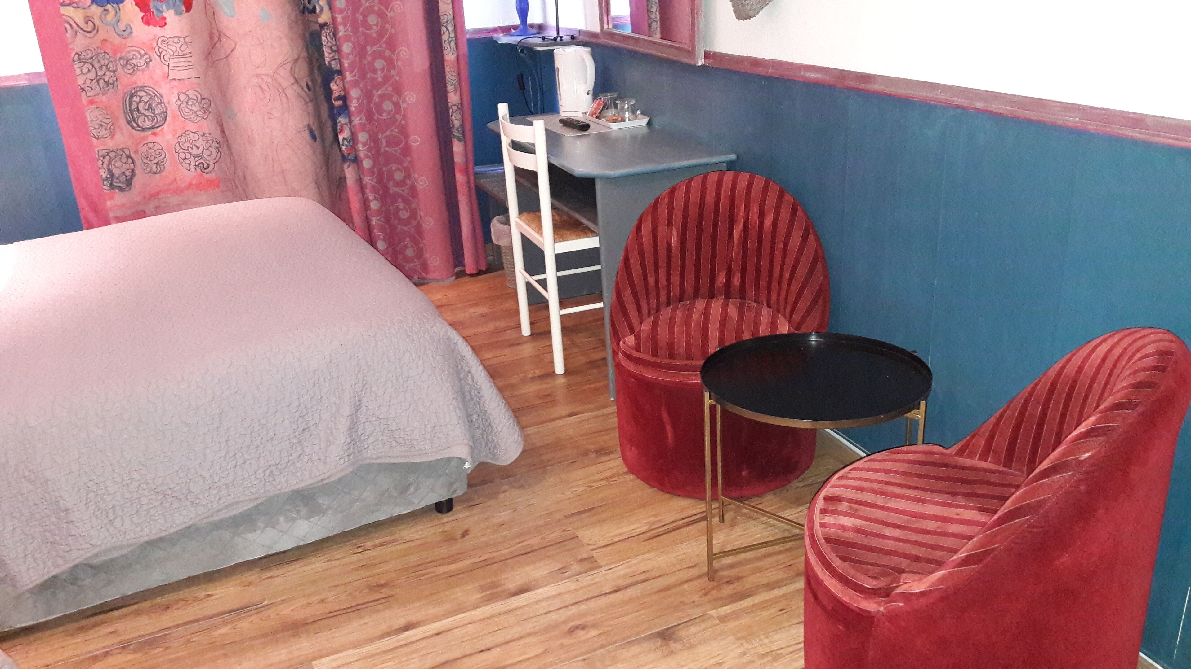 Een kamer met een bed, twee rode stoelen, een kleine tafel en een bureau met een waterkoker en kopjes.