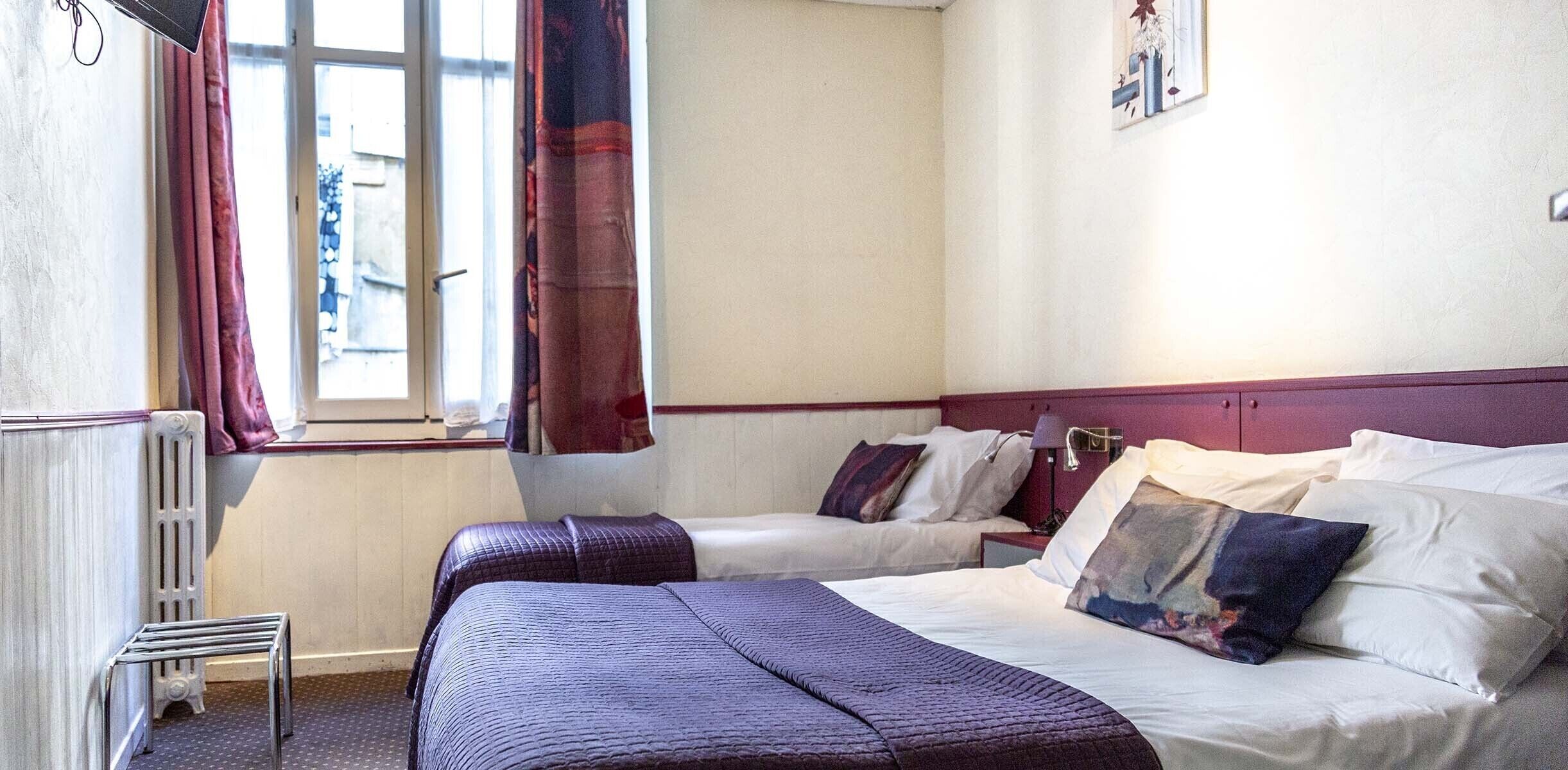 A hotel room with two beds, a radiator, a window with curtains, and a wall-mounted lamp.