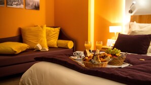 A hotel room with a bed, a bedside table with a lamp, a wall with framed pictures, and a breakfast tray with bread, fruit, and juice. - Hôtel Les Vallées (La Bresse)