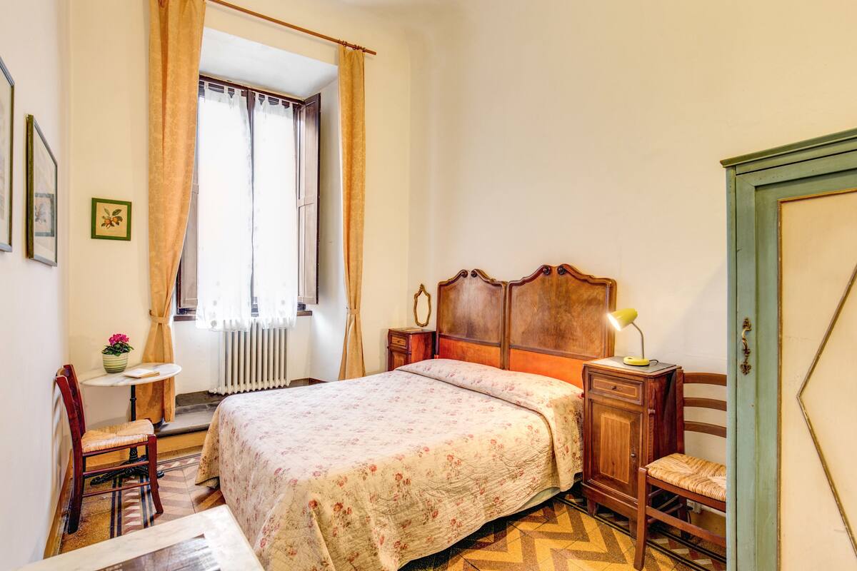 A bedroom with a wooden bed, a nightstand, a chair, a small table, and a window with curtains.