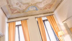 Una habitación con techo alto que presenta un fresco decorativo, dos ventanas con cortinas naranjas y una lámpara a la derecha.