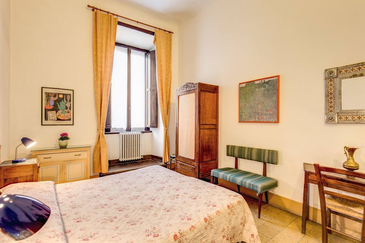A bedroom with a bed, a wooden wardrobe, a bench, a desk, and a mirror.