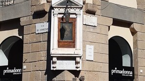 Fachada de edificio con un ventanal central que muestra una obra de arte enmarcada, flanqueado por dos escaparates con letreros de 'primadonna collection'.