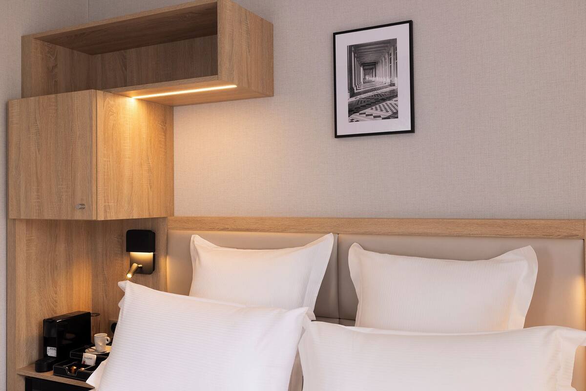 A neatly made bed with white linens, a wooden headboard, a wall-mounted light, and a framed picture above the bed.