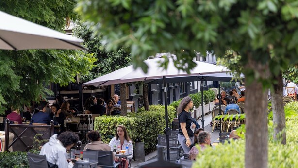 Un café al aire libre con mesas y sillas, rodeado de vegetación y sombrillas.