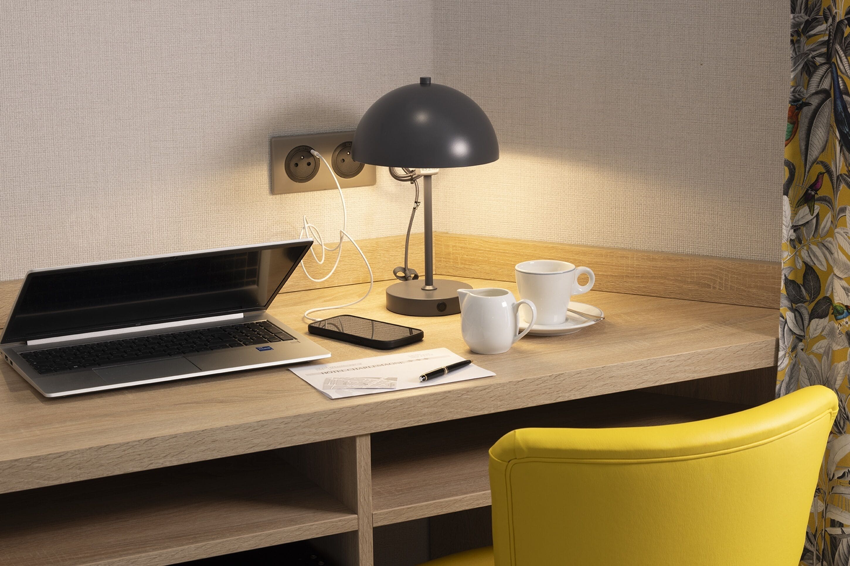 A wooden desk with a laptop, a smartphone, a white mug, and a black desk lamp.