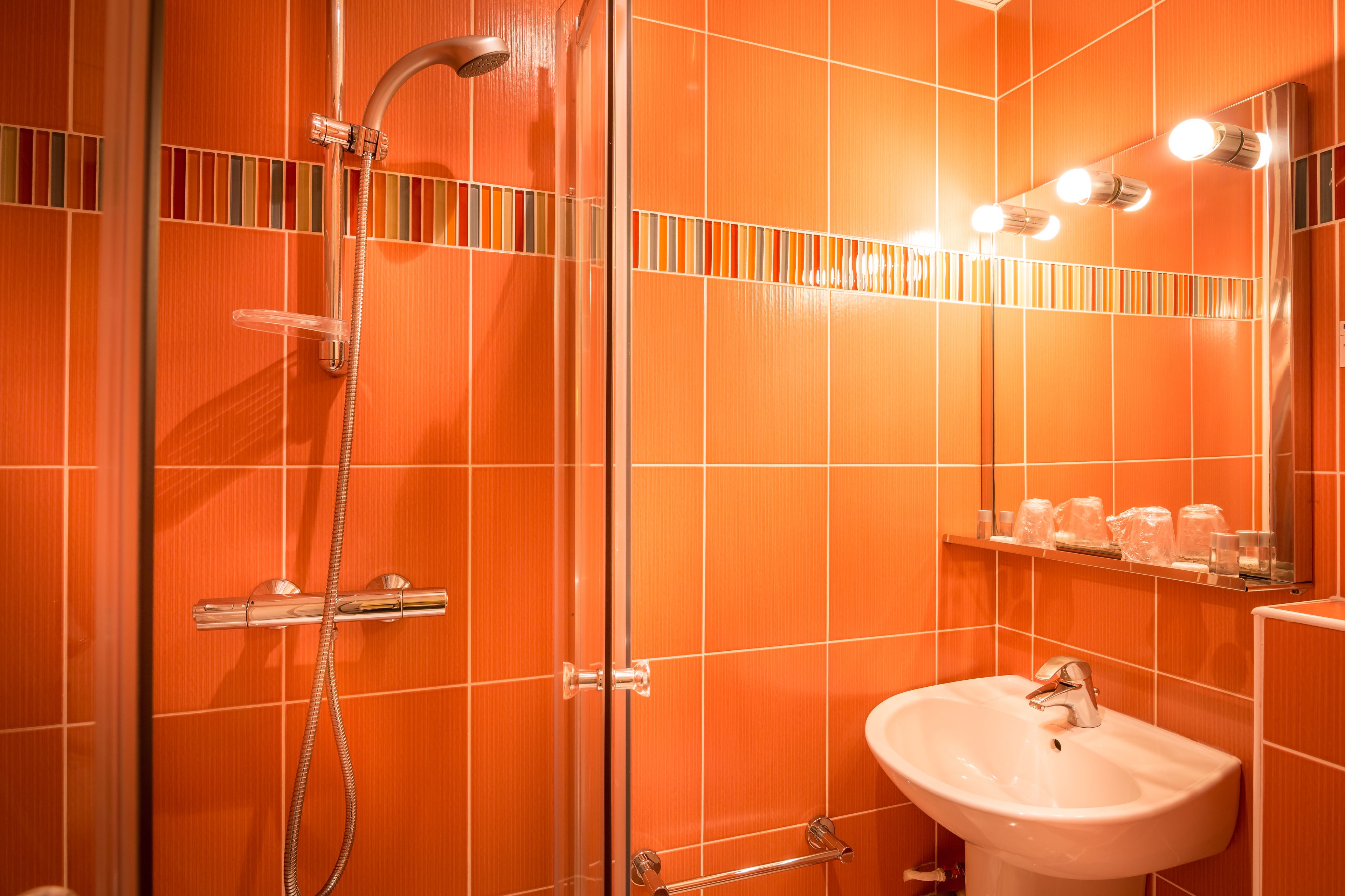 A bathroom with orange tiled walls, a glass shower enclosure, a white sink, and a mirror with lights.