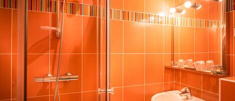 A bathroom with orange tiled walls, a glass shower enclosure, a white sink, and a mirror with lights.