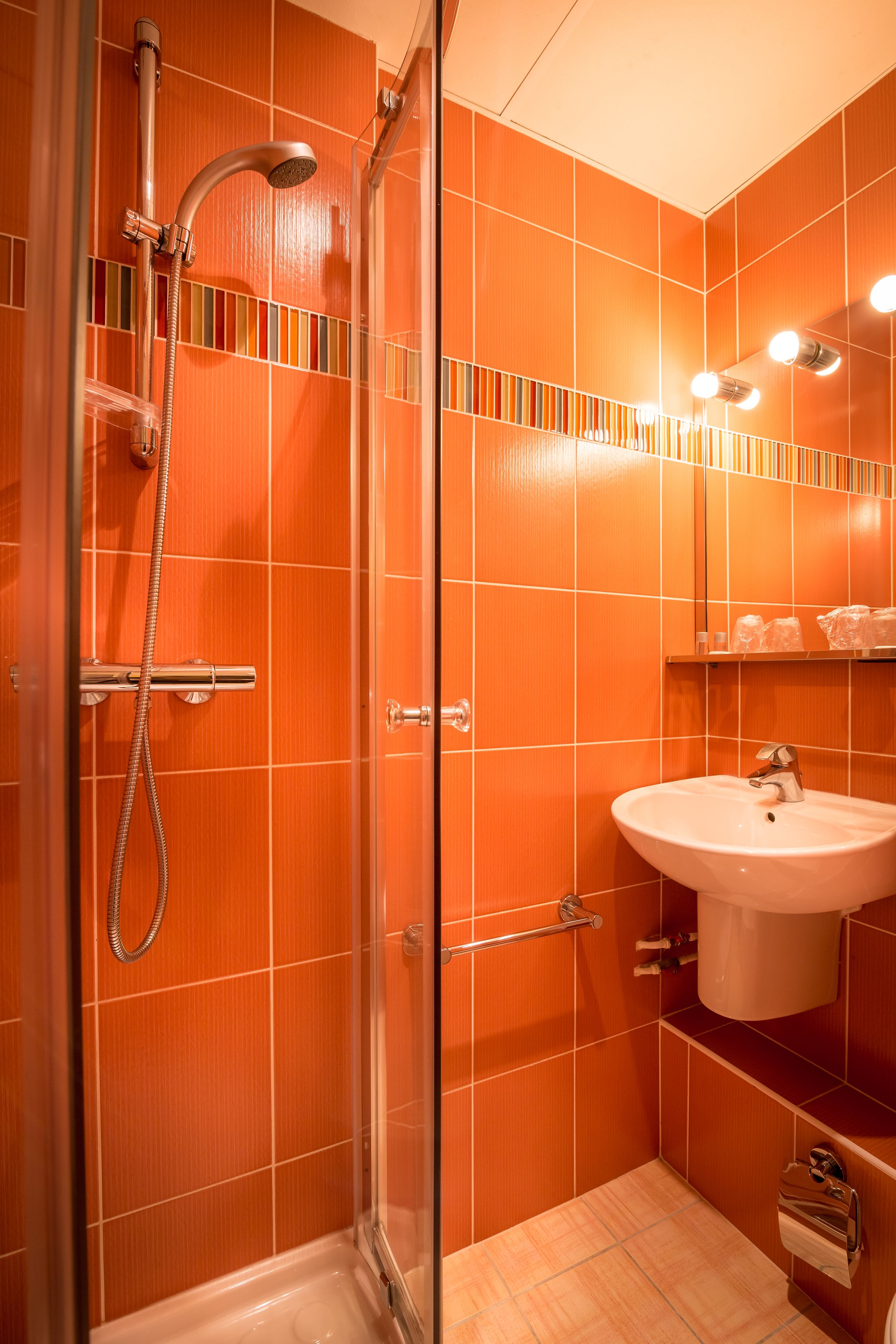 Um banheiro com azulejos laranja, chuveiro com divisória de vidro, pia branca e espelho iluminado.