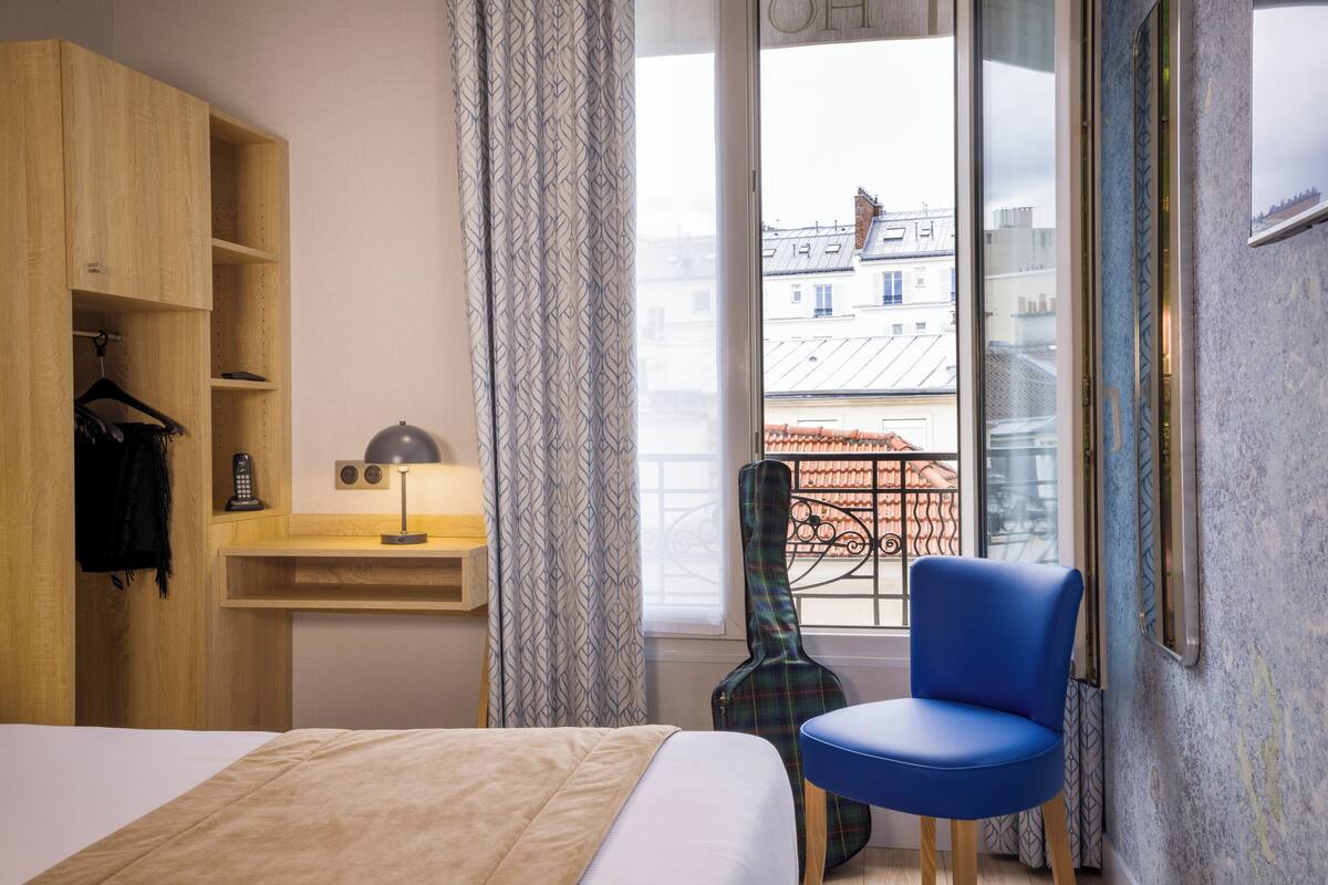 A hotel room with a bed, a wooden wardrobe, a desk with a lamp, a guitar, a blue chair, and a view of buildings through a window.