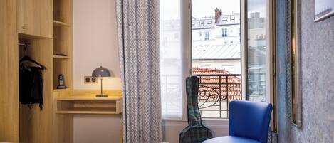 A hotel room with a bed, a wooden wardrobe, a desk with a lamp, a guitar, a blue chair, and a view of buildings through a window.