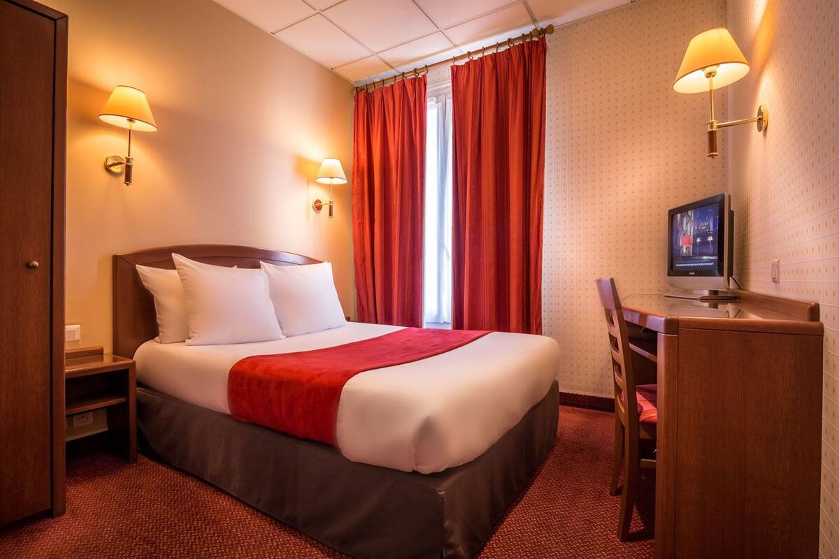 A hotel room with a bed, a desk, a television, and two wall-mounted lamps.