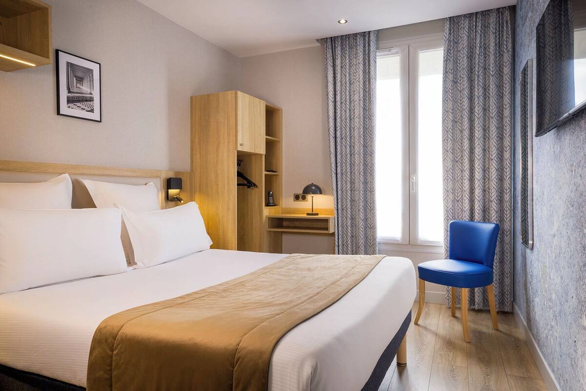 A hotel room with a large bed, a wooden wardrobe, a desk with a lamp, a blue chair, and a TV mounted on the wall.