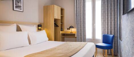 A hotel room with a large bed, a wooden wardrobe, a desk with a lamp, a blue chair, and a TV mounted on the wall.