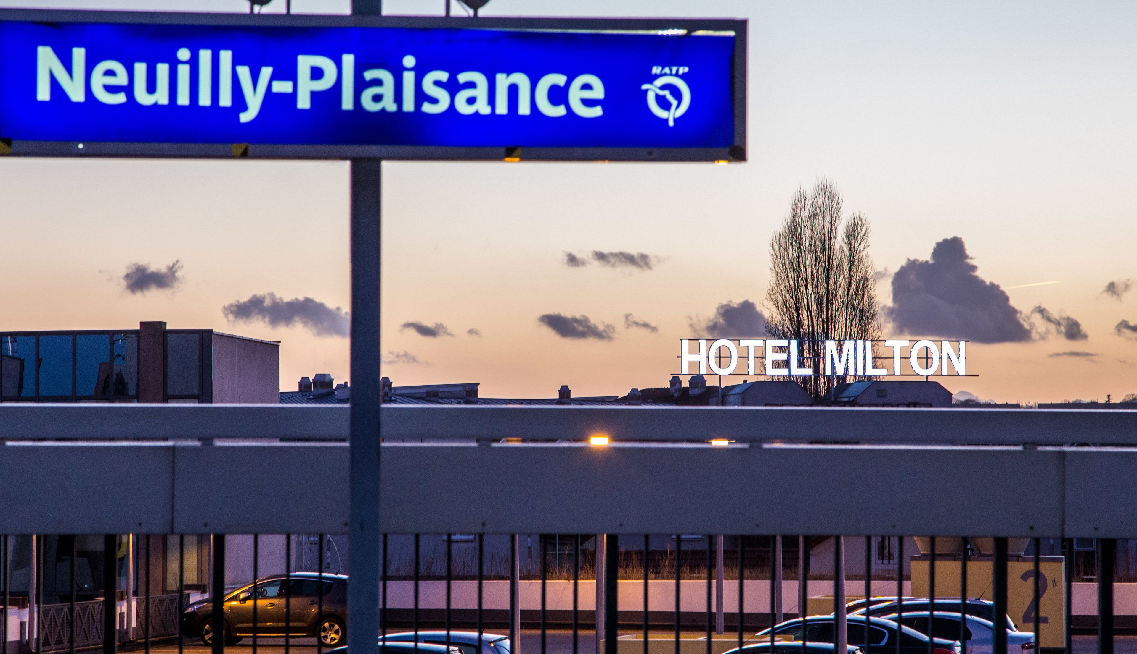 Une enseigne d’hôtel avec les inscriptions « Neully-Plaissance » et « HÔTEL MILTON », un espace de stationnement et un ciel au coucher du soleil.